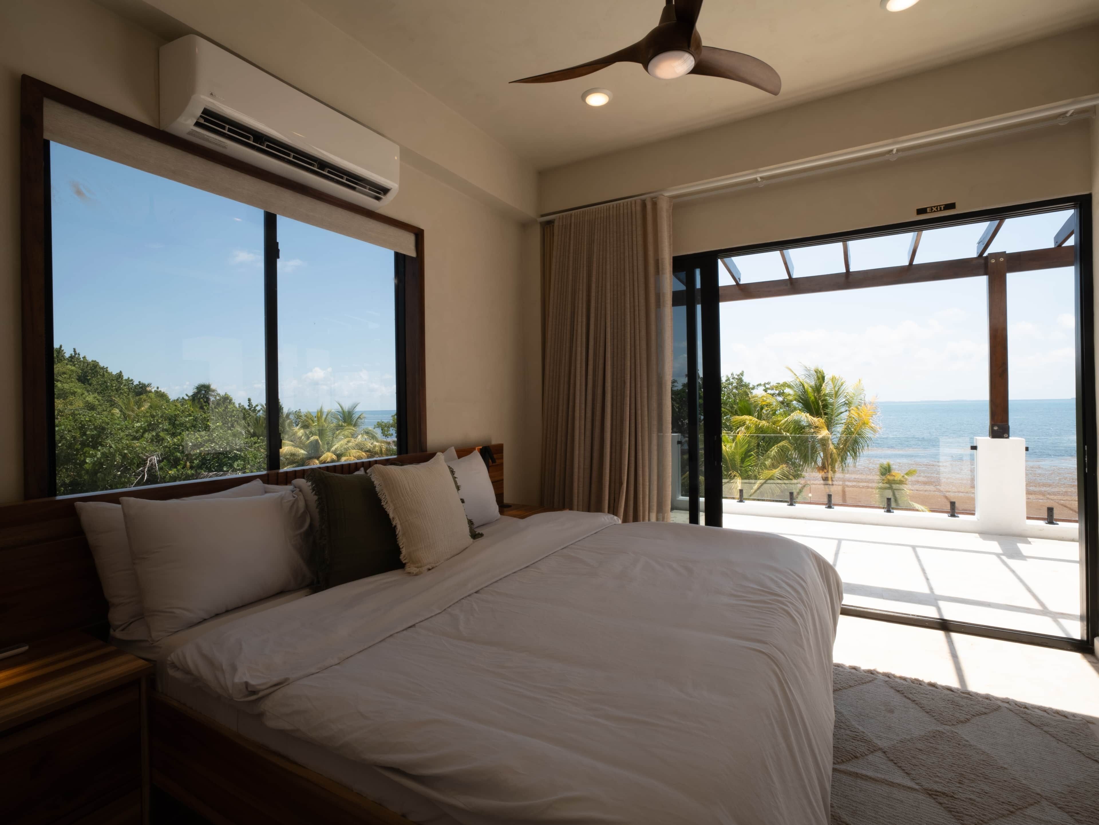 Un dormitorio con una cama grande, un equipo de aire acondicionado, un ventilador de techo y vistas al océano a través de ventanas amplias.