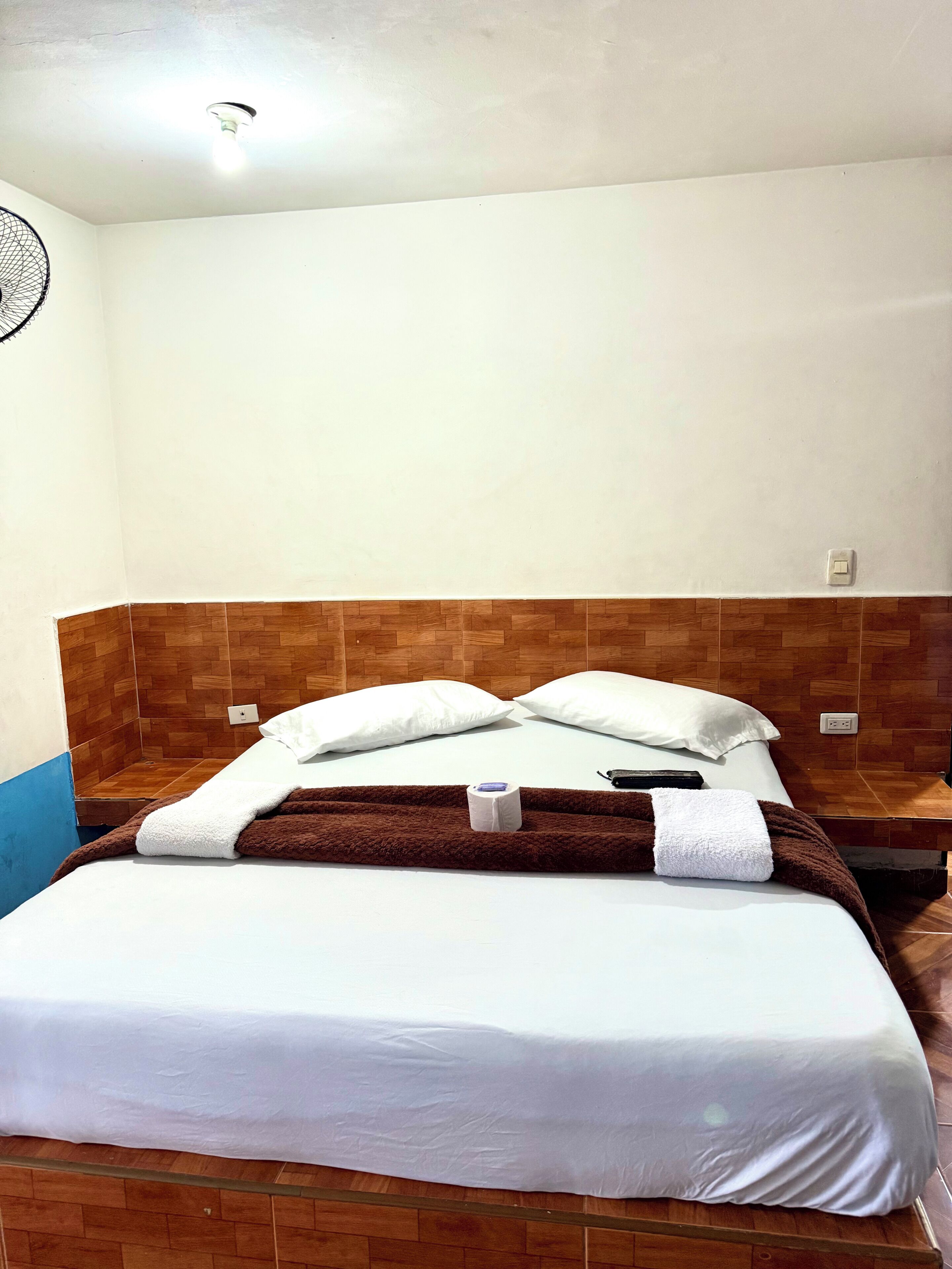 Quarto de hotel com cama, cabeceira de madeira, roupa de cama branca e uma luminária.
