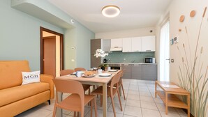 A modern kitchen with white cabinets, a dining table with chairs, a sofa with a pillow, and a small wooden table. - My Peschiera Holiday Apartment- Quadrivium 4 (Castelnuovo del Garda)