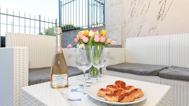 A wicker sofa, a table with a bottle of wine, glasses, a plate of pastries, and a vase of tulips. - My Peschiera Holiday Apartment- Quadrivium 4 (Castelnuovo del Garda)