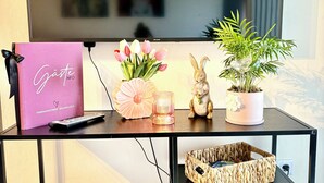A TV stand with a flat-screen TV, a pink bag, a vase with flowers, a candle, and a small rabbit figurine. - Vacation apartment Seelenglück with garden 🪴🦮 (Bad Harzburg)