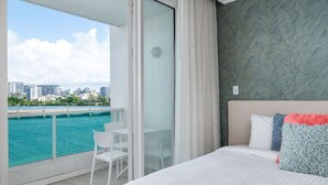 A hotel room with a balcony, a bed with pillows, a chair, and a view of a cityscape and water. - Lagoon Studio · Pool & Hot Tub (San Juan)