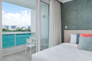 A hotel room with a balcony, a bed with pillows, a chair, and a view of a cityscape and water. - Lagoon Studio · Pool & Hot Tub (San Juan)