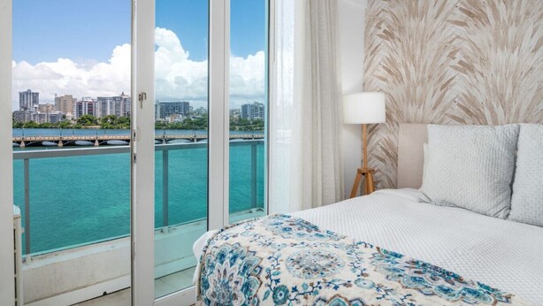 A hotel room with a bed, a nightstand, a lamp, and a view of the water and city skyline. - Waterfront Studio · Pool · Water Backup (San Juan)