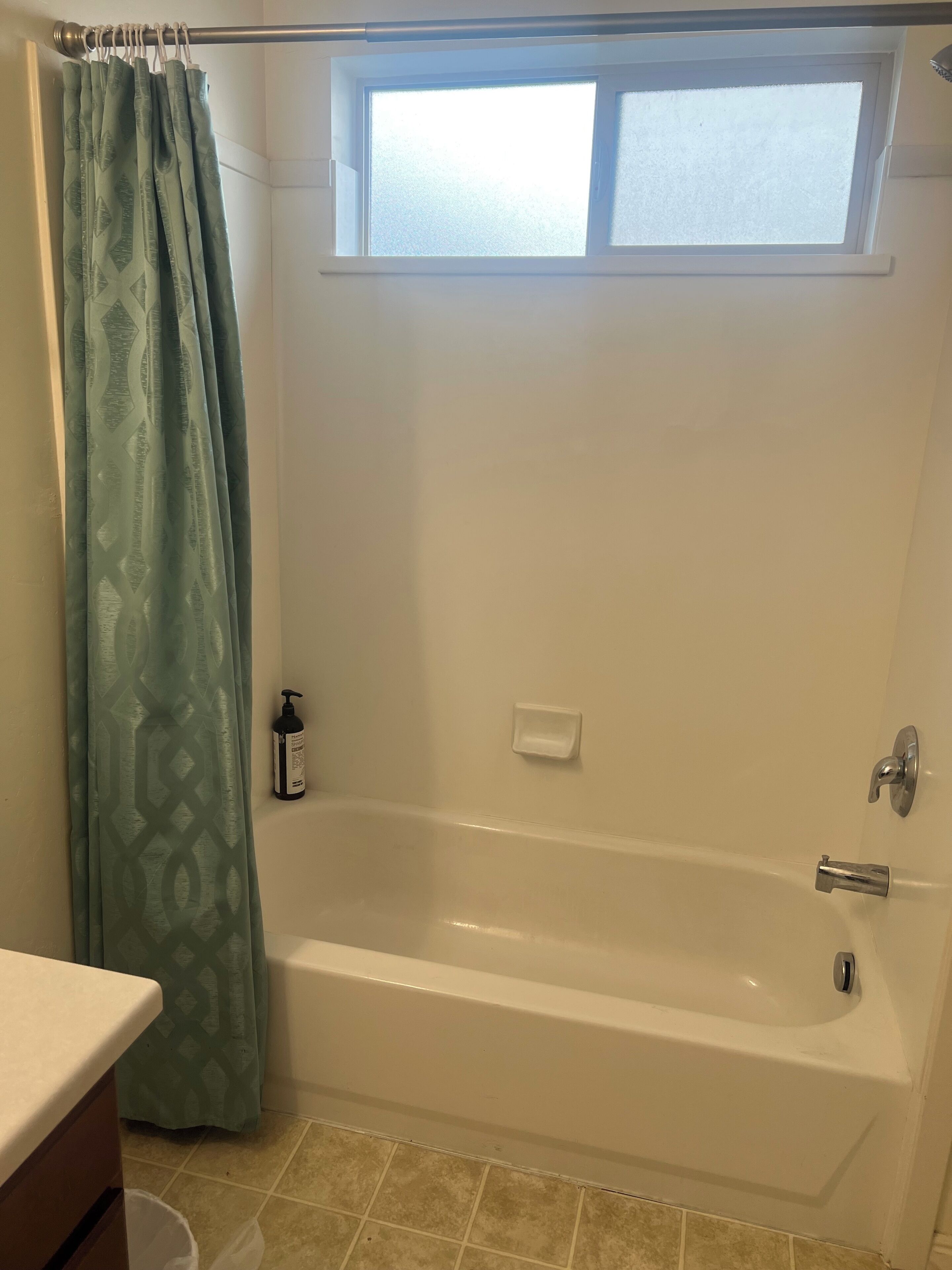 A bathroom with a bathtub, a window, a shower curtain, a soap dispenser, and a trash bin.
