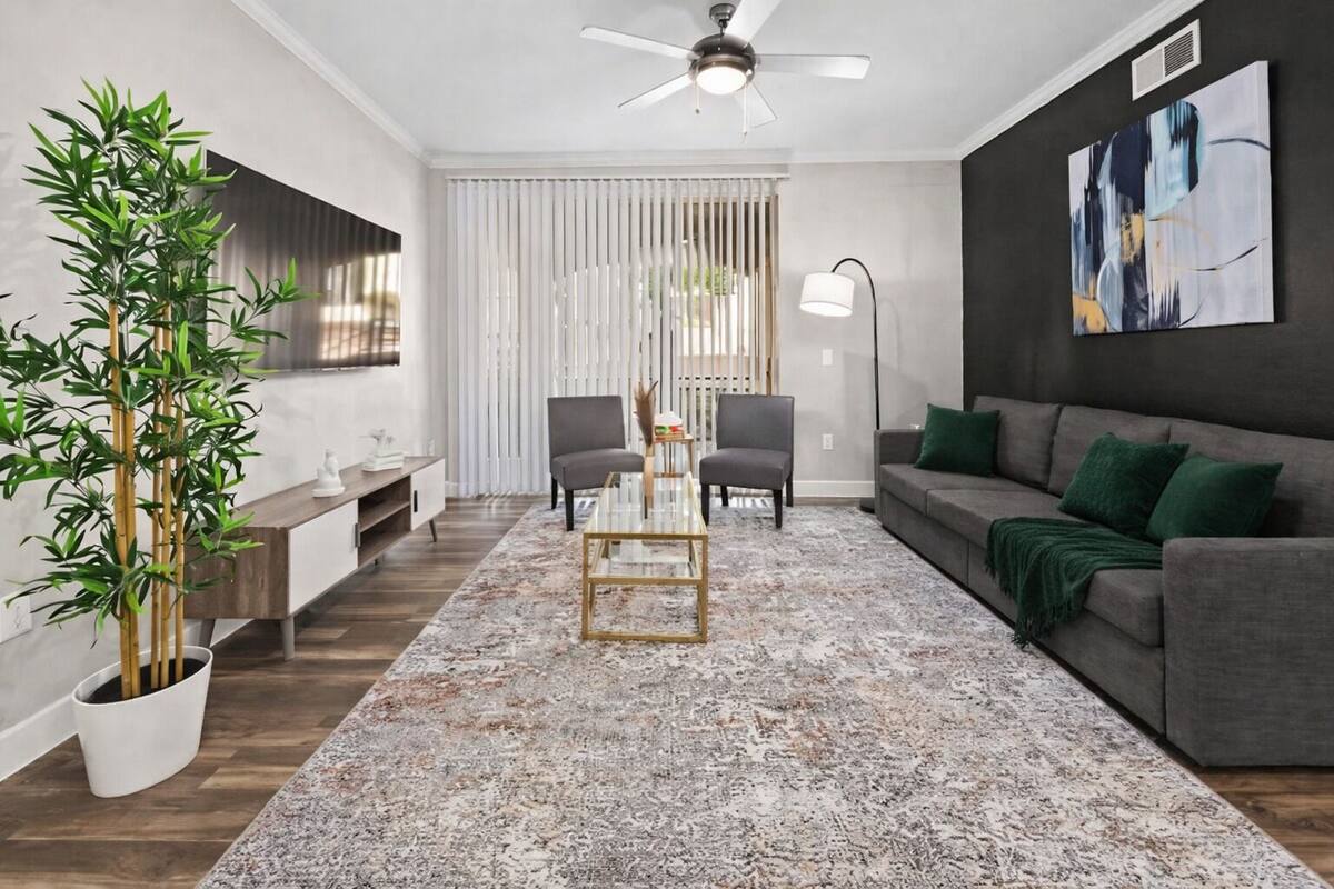Una sala de estar moderna con una mesa de centro de vidrio, dos sillones, un sofá, un cuadro abstracto grande, una planta de bambú y un ventilador de techo.