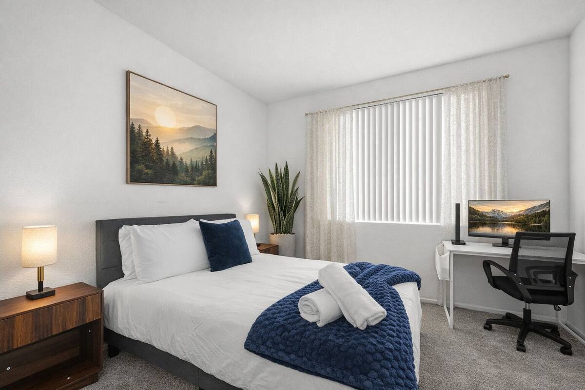 Habitación de hotel con cama, escritorio, silla, planta y ventana con persianas.