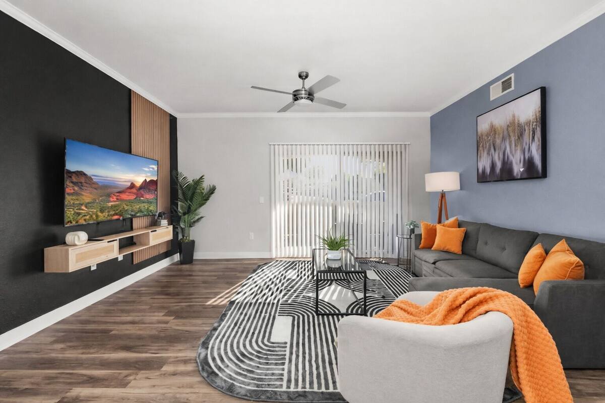 Una sala de estar moderna con un televisor de pantalla plana, un sofá gris con cojines anaranjados, una alfombra estampada y un ventilador de techo.