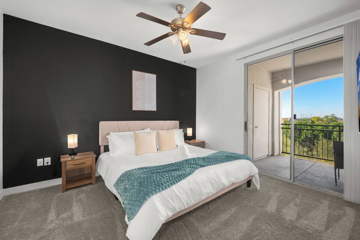 Un dormitorio con cama, mesitas de noche, ventilador de techo, ventana grande y balcón.