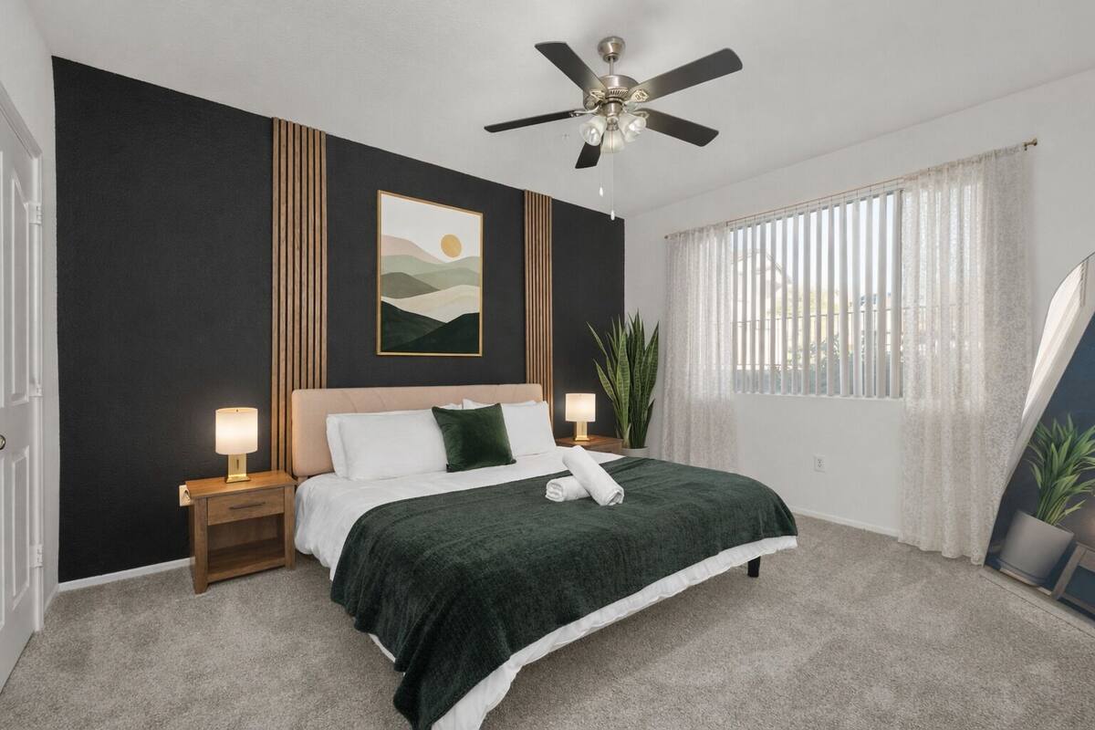 Un dormitorio con cama, lámparas de noche, ventilador de techo, ventana con cortinas y un cuadro colgado en la pared.