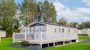 A single-story mobile home with a covered porch and a small deck, surrounded by green grass and trees. - Seton Sands 3BR Premium Caravan - Near Edinburgh (Port Seton)