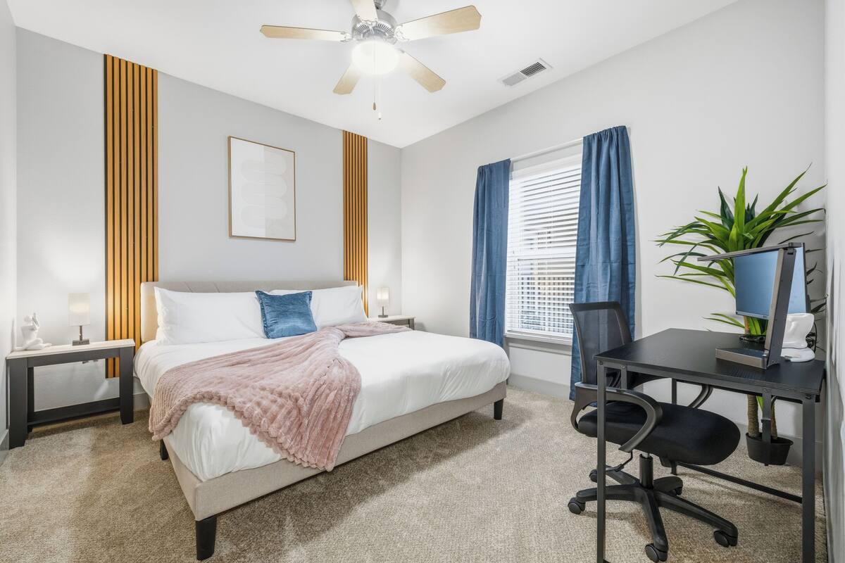A bedroom with a bed, a desk, a chair, a ceiling fan, a window with curtains, and a plant.