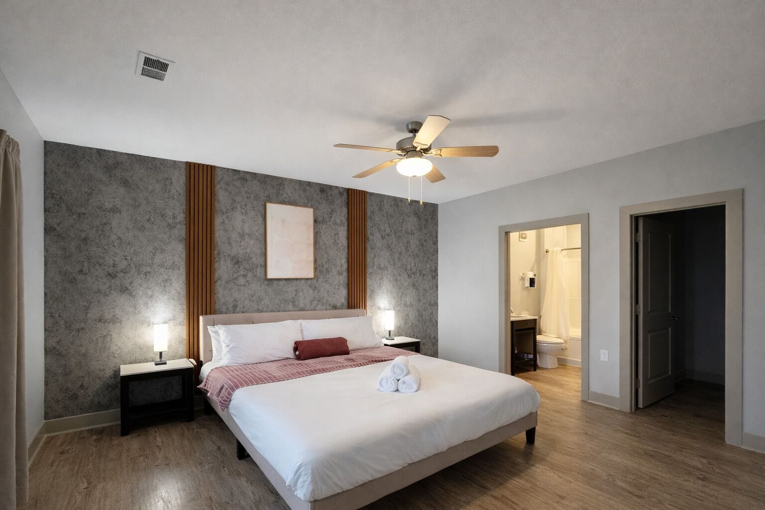 Un dormitorio con cama, lámparas de noche, ventilador de techo, un cuadro enmarcado y un baño visible a través de una puerta abierta.