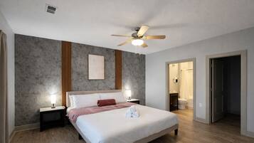 Un dormitorio con cama, lámparas de noche, ventilador de techo, un cuadro enmarcado y un baño visible a través de una puerta abierta.
