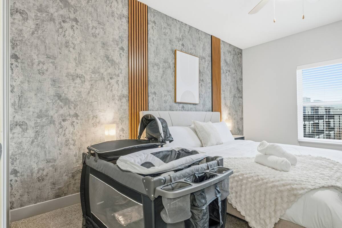 A hotel room with a bed, a crib, a window with blinds, and a city view.