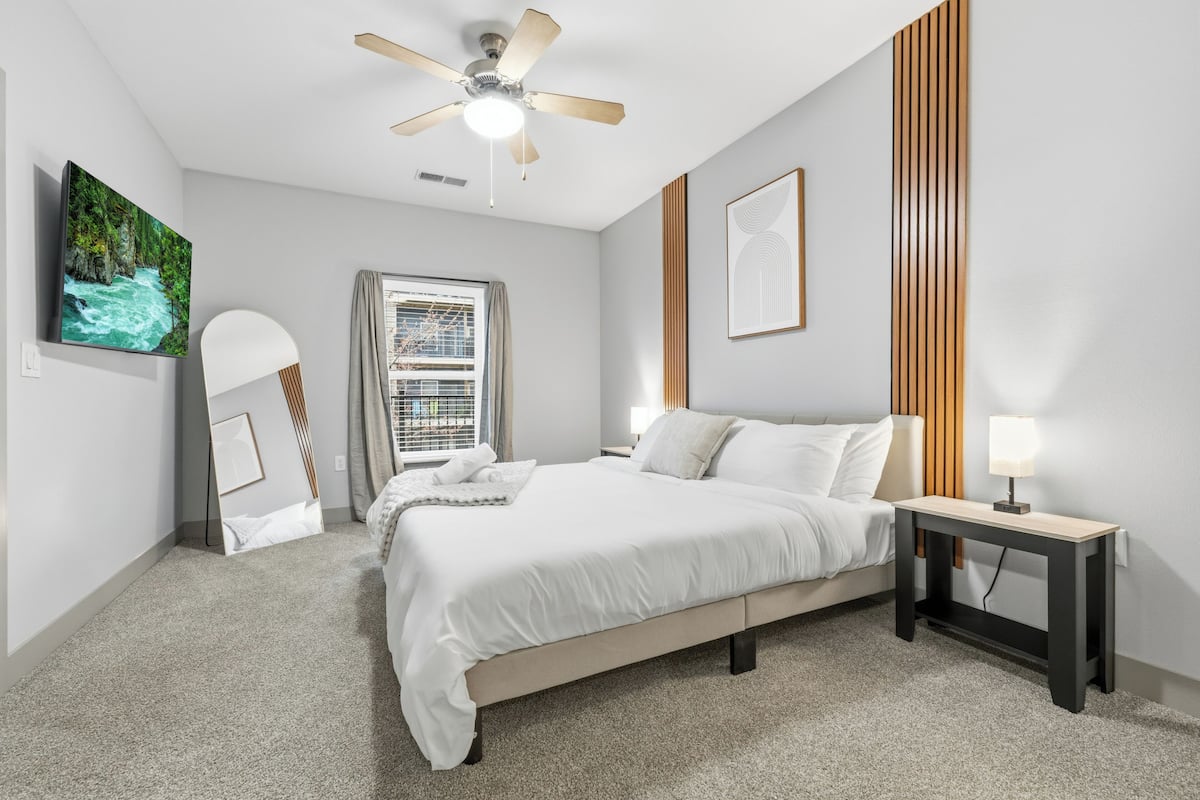 A bedroom with a bed, a bedside table with a lamp, a ceiling fan, a window with blinds, and a painting on the wall.