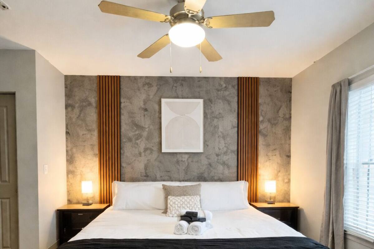A hotel room with a large bed, a ceiling fan, two bedside lamps, a framed artwork on the wall, and a window with curtains.