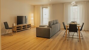 Een moderne woonkamer met een flatscreen televisie, een grijze bank, een eettafel met stoelen en een houten vloer.
