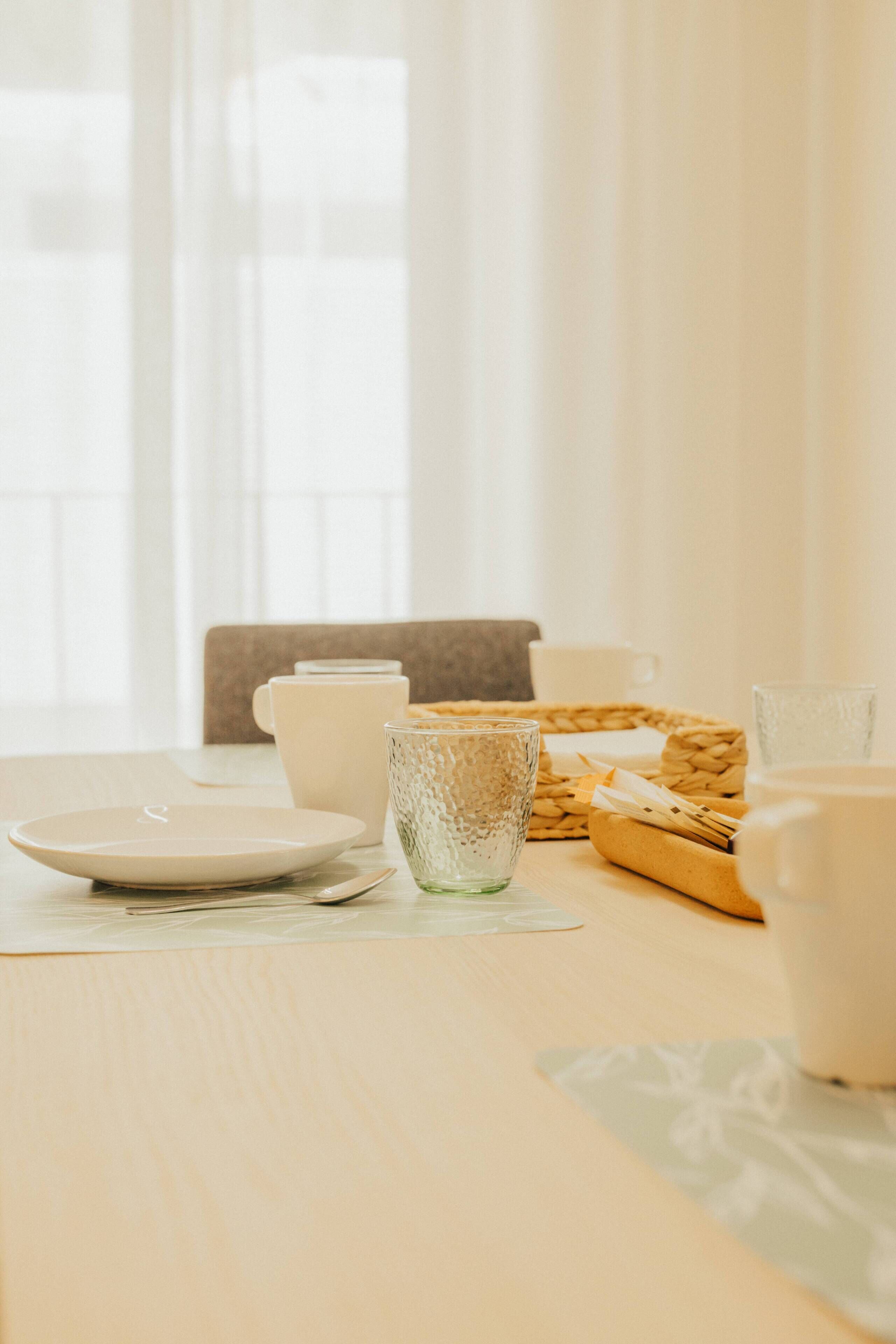 Una mesa de comedor con platos, tazas y un vaso, frente a una ventana con cortinas translúcidas.