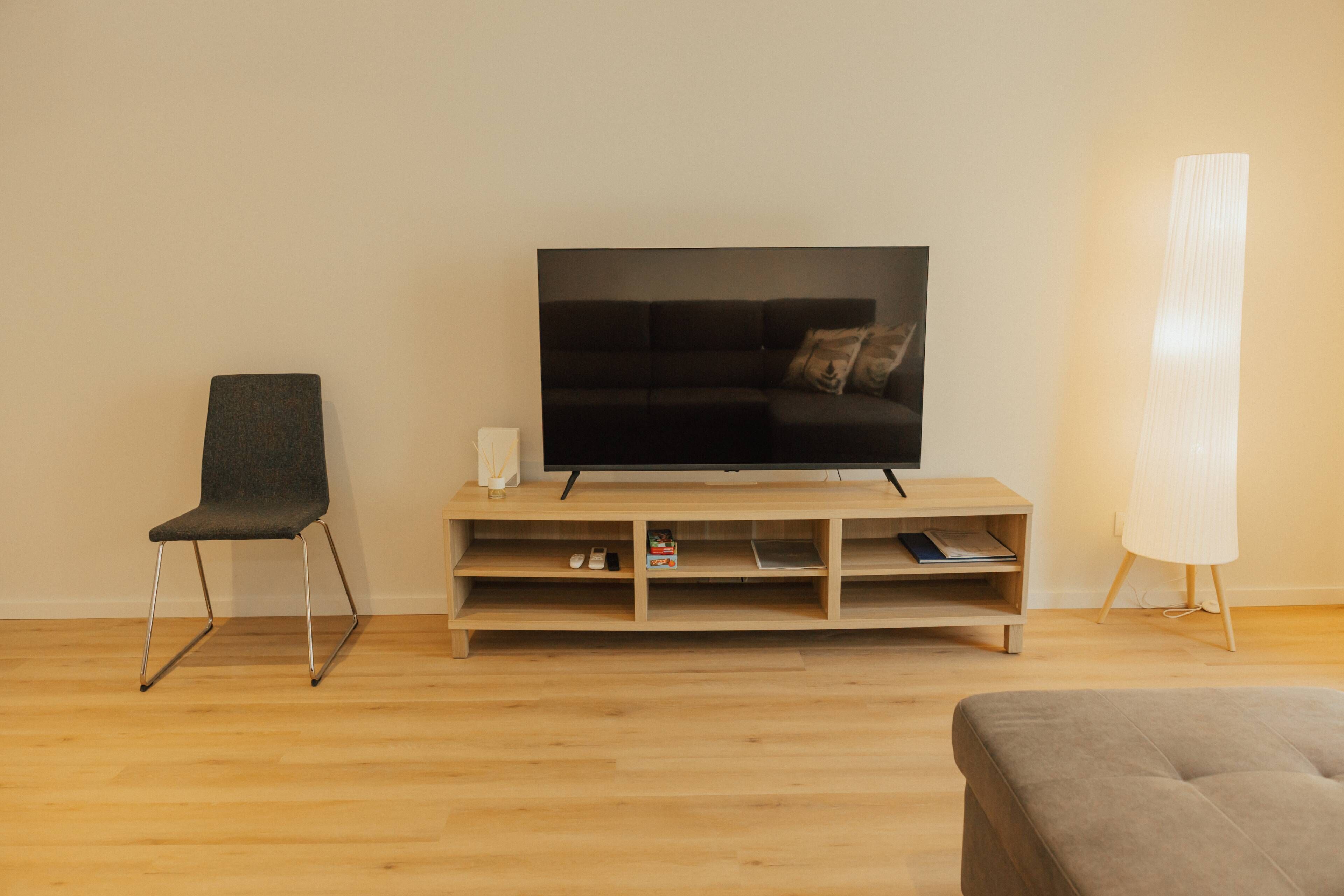 Un salon moderne avec un téléviseur à écran plat posé sur un meuble en bois, un fauteuil gris, un pouf gris et une lampe sur pied.