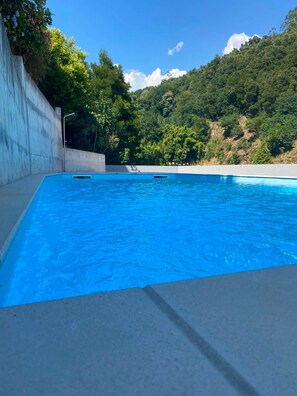 A swimming pool with a concrete edge, surrounded by greenery and a hillside.