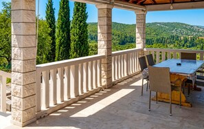 A covered outdoor area with stone pillars, a table with chairs, and a view of green hills.