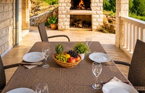 An outdoor dining area with a stone fireplace, wicker chairs, a table set with plates, glasses, and a fruit bowl.