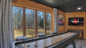Ein Zimmer mit Shuffleboard-Tisch, großen Fenstern mit Blick auf den Wald, einem Netflix-Logo an der Wand und einer Holzbalkendecke.