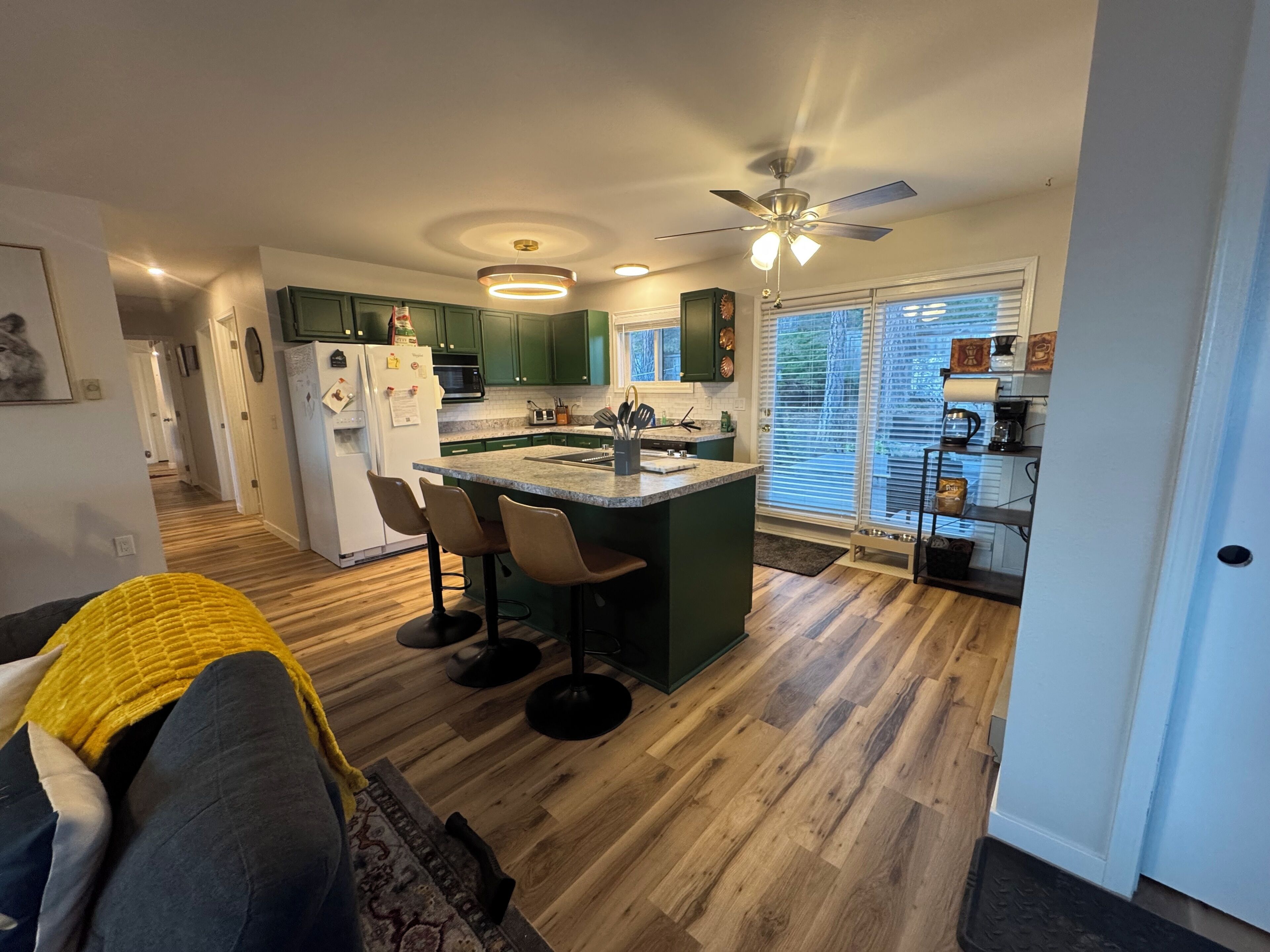 A modern kitchen with green cabinets, a central island with bar stools, a refrigerator, and a ceiling fan.