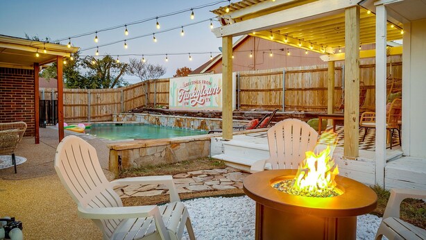 Ein Garten mit Pool, Adirondack-Stühlen, einer Feuerstelle und einem Pergola mit Lichterketten.