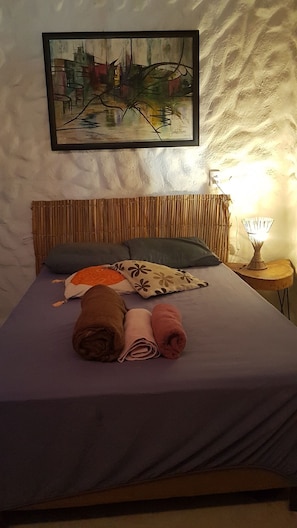 A bed with a purple bedspread, two rolled towels, a pillow, and a framed abstract painting on the wall. - Charming and private house near the best beach in Porto Seguro. (Porto Seguro)