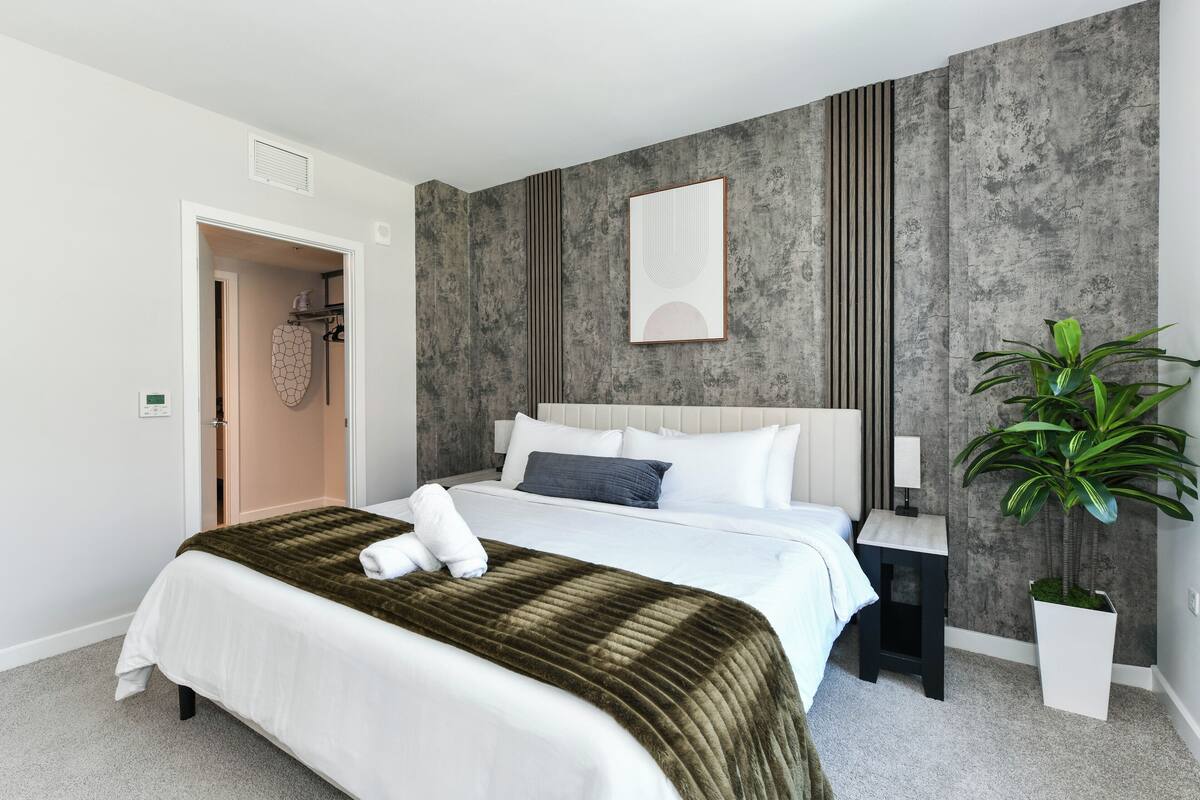 Une chambre moderne avec un grand lit, une table de chevet, une commode, une œuvre d’art accrochée au mur, une plante en pot et une porte donnant sur une salle de bain.