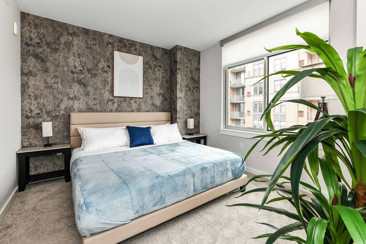 Une chambre à coucher comprenant un lit, une table de chevet, un tableau décoratif au mur, une fenêtre et une plante en pot.