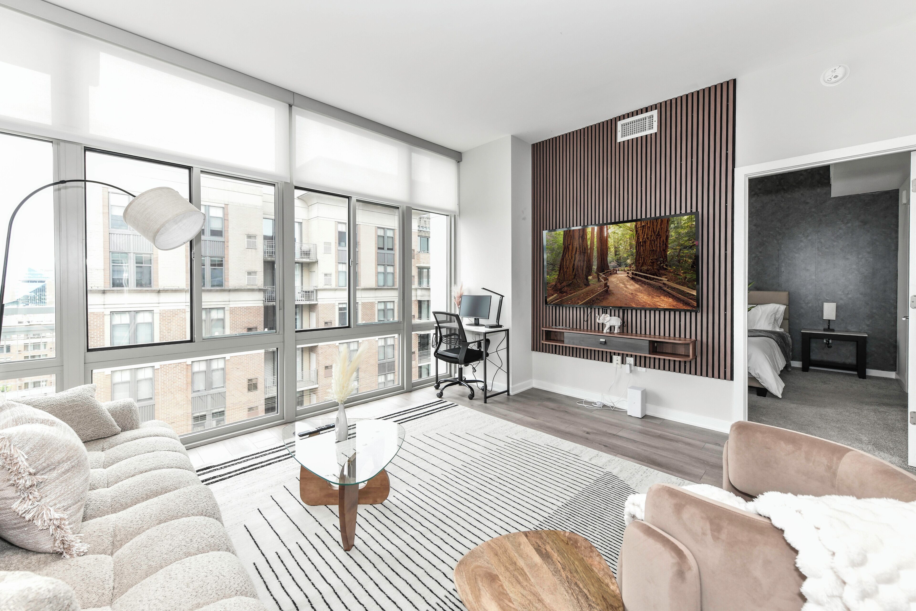 Una habitación de hotel moderna con ventanas amplias, un televisor de pantalla plana, un sofá, una mesa de centro y un escritorio con una silla.