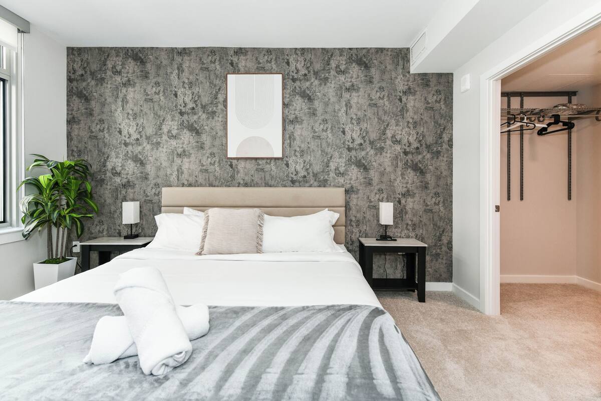 Une chambre moderne avec un grand lit, des tables de chevet, une œuvre d’art accrochée au mur, une plante en pot et une armoire contenant des vêtements.