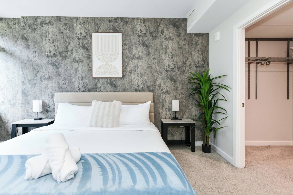 Une chambre à coucher moderne avec un grand lit, deux tables de chevet avec des lampes, un mur gris à texture et une plante en pot.