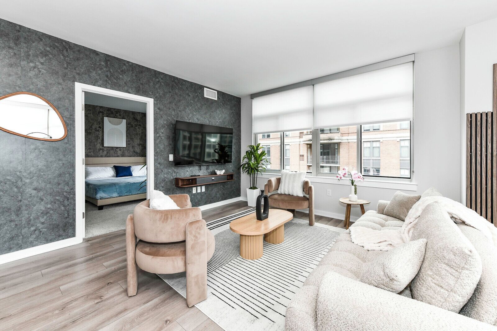 A modern living room with a grey wall, a flat-screen TV, a sofa, a coffee table, and a view of the city through large windows.