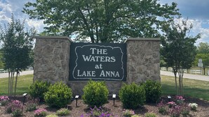 Señalización de 'The Waters at Lake Anna' con un muro de piedra, un árbol grande y macizos de flores ajardinados.