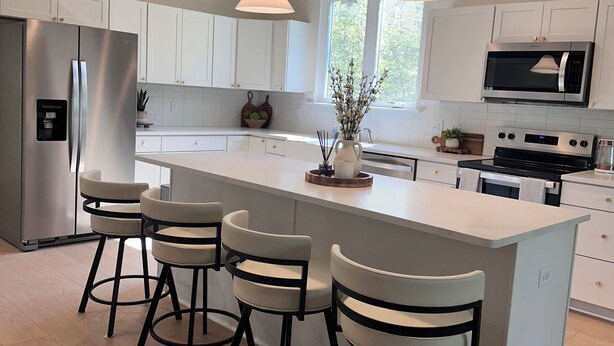 Una cocina moderna con una isla central, taburetes de bar, electrodomésticos de acero inoxidable, iluminación colgante y armarios blancos.