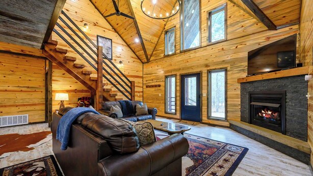 A log cabin interior with a fireplace, leather sofa, and a staircase. - Drift Inn, amazing back yard privacy! hot tub (Broken Bow)