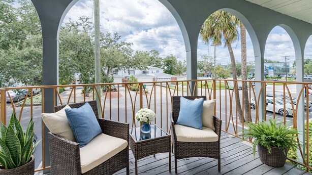 A covered outdoor area with wicker furniture, potted plants, and a view of a street and buildings.