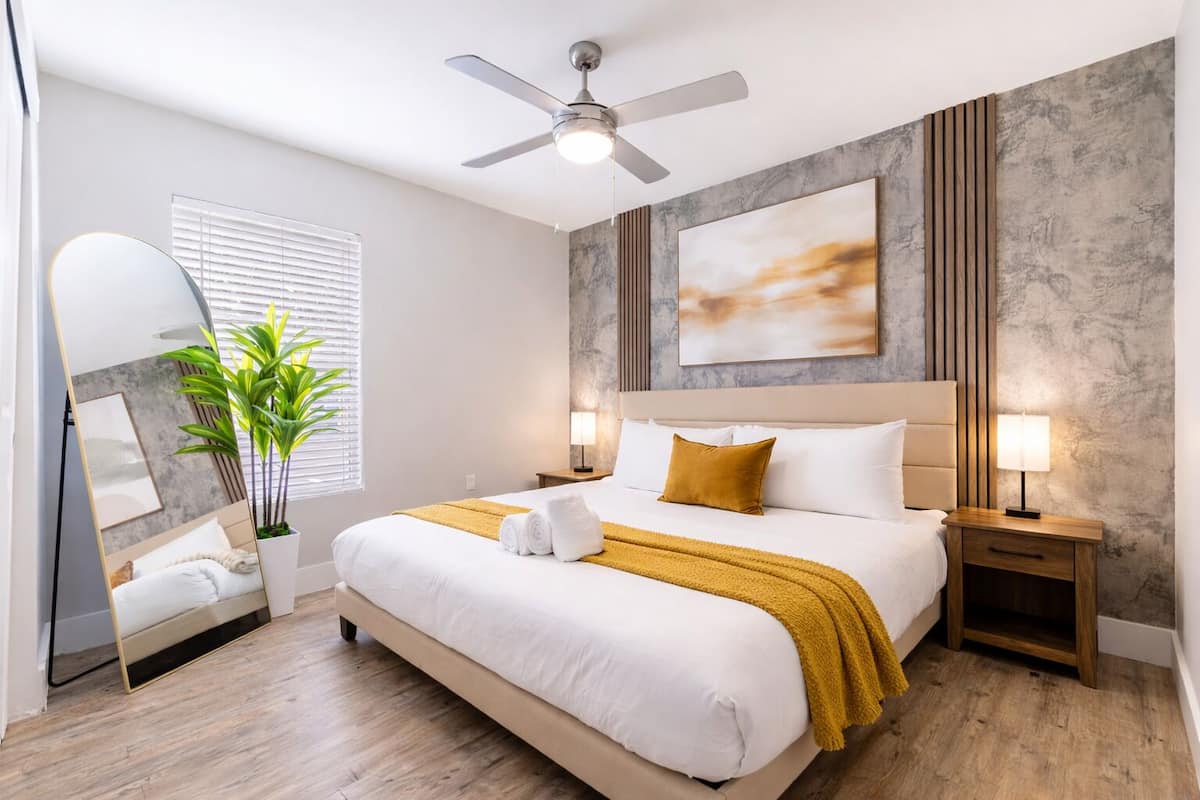 Habitación de hotel con cama, lámparas de noche, ventilador de techo, espejo grande y una planta.