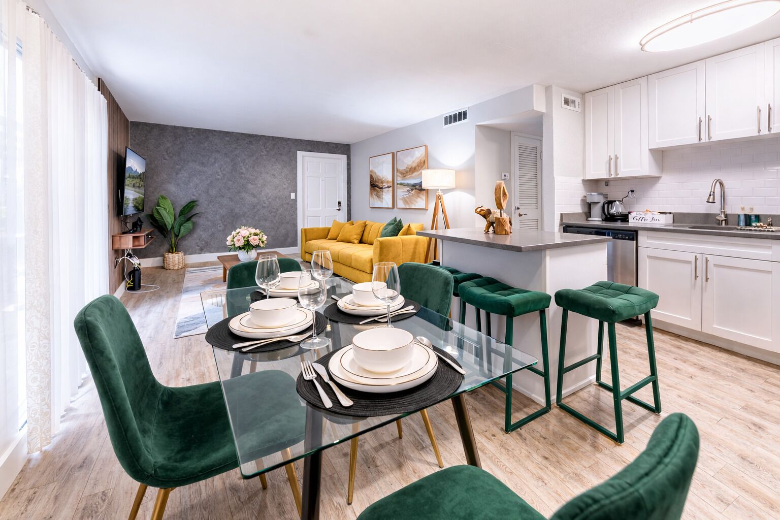 A modern kitchen with white cabinets, a dining area with a glass table and green chairs, and a yellow sofa in the background.