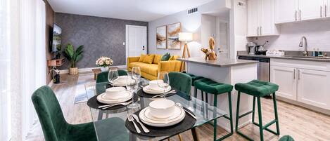 A modern kitchen with white cabinets, a dining area with a glass table and green chairs, and a yellow sofa in the background.