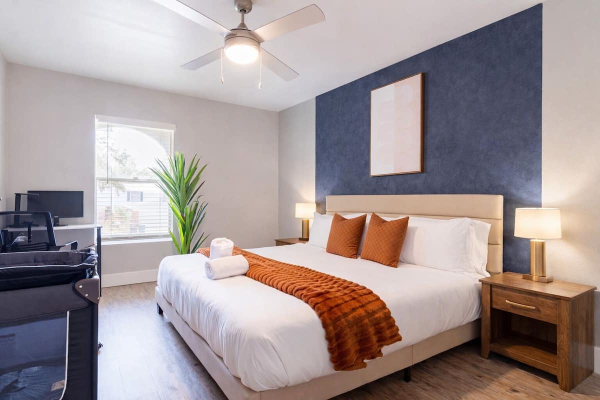 Habitación de hotel con cama, mesitas de noche, escritorio, silla, planta y ventilador de techo.