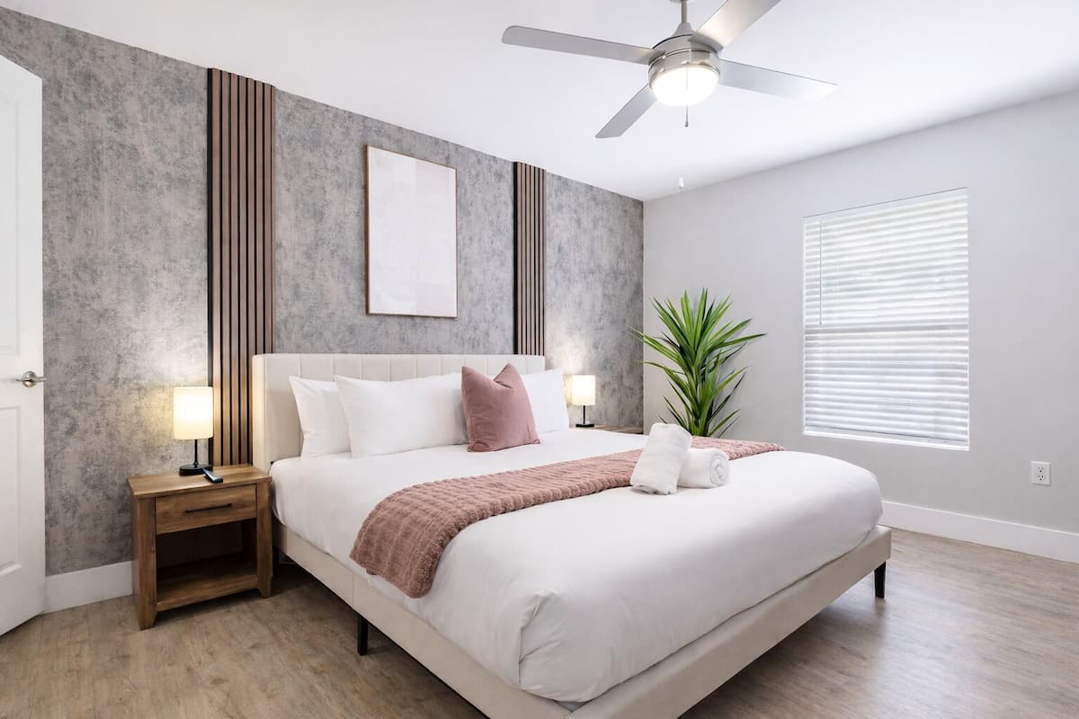 Un dormitorio moderno con cama, lámparas de noche, ventilador de techo, planta en maceta y ventana con persianas.
