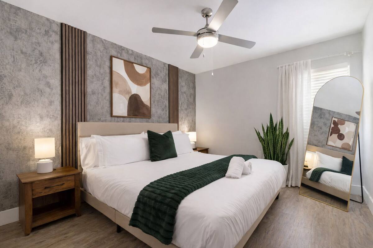 Habitación de hotel con cama, mesitas de noche, ventilador de techo, un espejo grande y una planta en maceta.