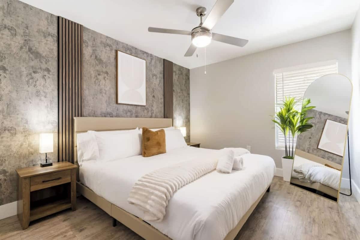 Un dormitorio con cama, mesitas de noche, ventilador de techo, ventana con persianas y una planta en maceta.