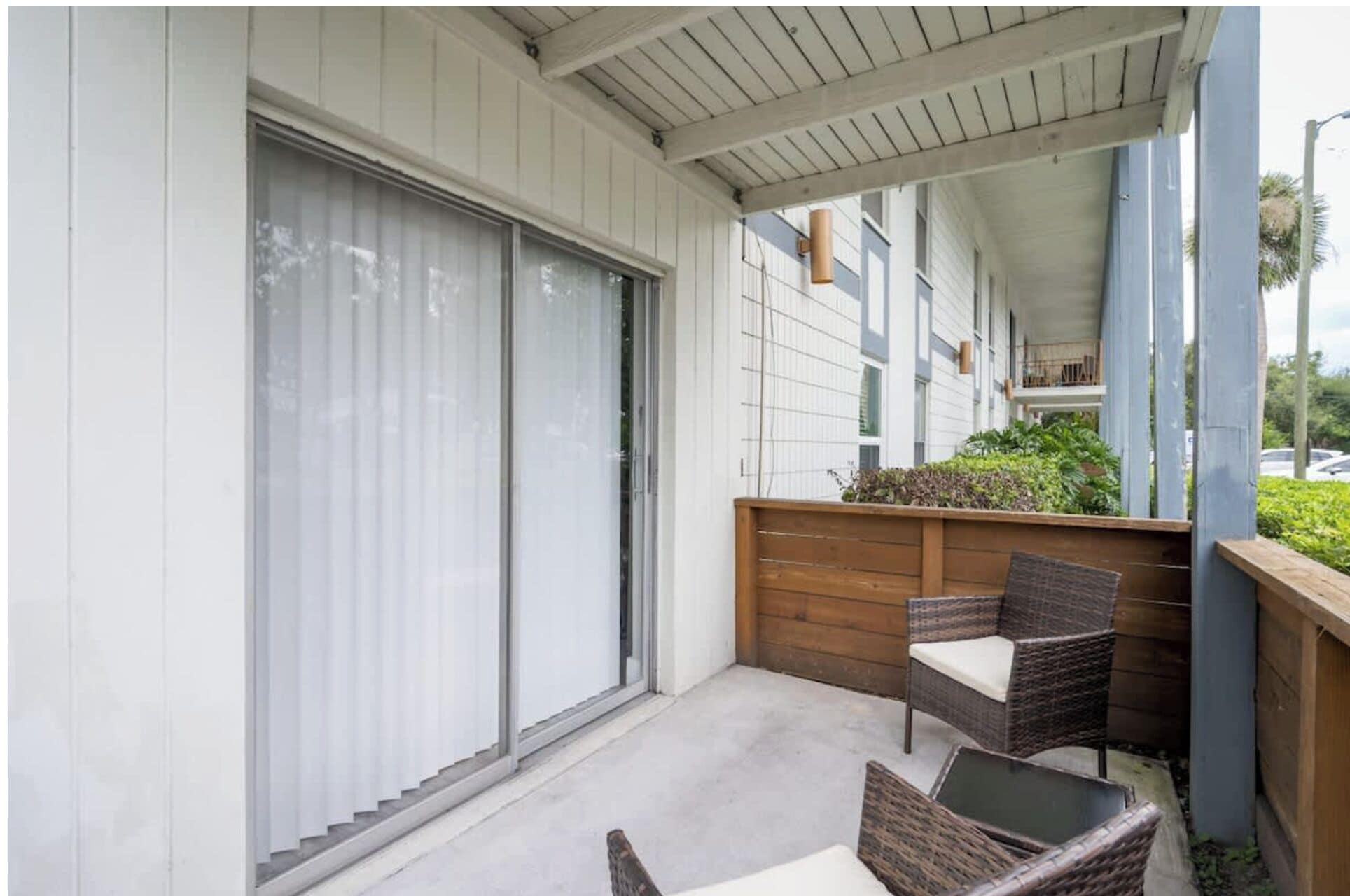 A covered balcony with a sliding door, wicker furniture, and a view of a building with a balcony.