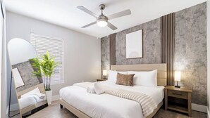 A modern bedroom with a bed, nightstands, a ceiling fan, a potted plant, and a large window with blinds.